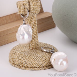 May include: A pair of silver hoop earrings with large, white baroque pearl drops. The earrings are displayed on a hessian stand.