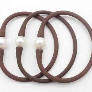 Pode incluir: Três pulseiras de silicone marrom com um detalhe de pérola branca em cada uma. As pulseiras estão empilhadas uma em cima da outra.
