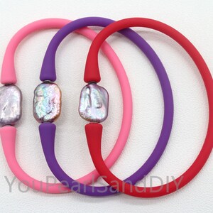 May include: Three colorful bracelets. Each bracelet features a large, iridescent, rectangular pearl bead. The bracelets are made of flexible, rubber-like material in pink, purple, and red. The beads are connected by a small, silver-colored metal piece.