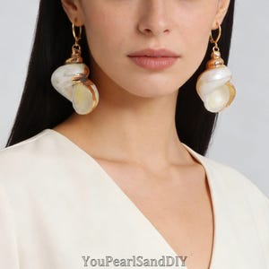 Peut inclure: Une paire de boucles d'oreilles créoles dorées avec de grands pendentifs en coquillage blanc irisé, ornés d'accents métalliques dorés sur le dessus. Le texte YouPearlSandDIY est visible en bas. Les boucles pendent à de fins anneaux dorés.