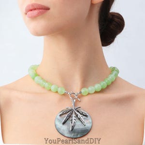 May include: A necklace featuring light green round beads and a silver-toned metal pendant shaped like a leaf, centred on a dark iridescent circular shell disc. The piece includes a silver-toned toggle clasp and the text YouPearlSandDIY.