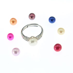 May include: A silver adjustable ring with a large white pearl center stone, surrounded by small clear stones. Scattered around the ring are seven colorful pearls in shades of pink, orange, purple, blue, and yellow.