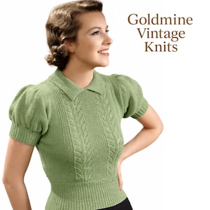 May include: A pale green knitted top with short, puffed sleeves and a collared neckline. The top has a central cable knit pattern and is labelled "Goldmine Vintage Knits". The woman is smiling and wearing the top.