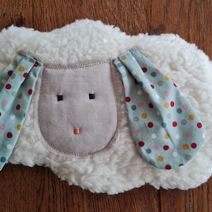 Könnte beinhalten: Ein weißes, flauschiges Stofftier in Form eines Schafs mit einem hellbraunen Gesicht und zwei Ohren mit grünem und weißem Punkte-Muster.