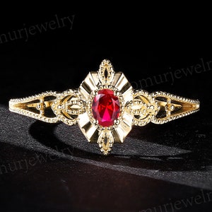 Puede incluir: Un anillo dorado con una gema roja ovalada en el centro. El anillo presenta un diseño de filigrana ornamentado alrededor de la gema y a lo largo de la banda. El anillo se encuentra sobre un fondo oscuro.