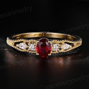 Könnte beinhalten: Ein goldener Ring mit einem ovalen, tiefroten Edelstein in der Mitte, flankiert von kleineren weißen Edelsteinen. Das Band weist aufwendige Details auf. Der Ring ist vor einem dunklen Hintergrund platziert, der sein Funkeln hervorhebt.