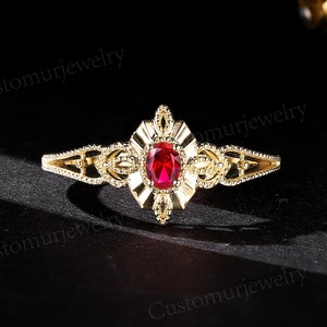 May include: A gold ring with an oval, red gemstone at the centre. The ring has detailed filigree work around the gemstone and along the band. The ring is set against a dark background, accentuating its design.