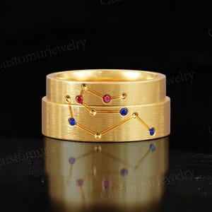 Peut inclure: Deux bagues dorées superposées, chacune ornée d'un motif de constellation avec des pierres précieuses rouges et bleues. Les bagues sont posées sur un fond noir. Le texte "Customurjewelry" est visible.