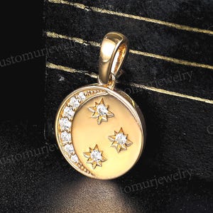 May include: Gold-tone pendant featuring a crescent moon and three stars. The moon is adorned with a row of small, clear gemstones. The stars are also embellished with clear gemstones. The pendant has a loop for a chain.
