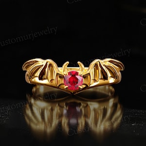 May include: A gold ring with a red gemstone in the center, shaped like a bat with wings spread out.