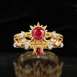 May include: A gold-colored ring with a large, round, red gemstone at the center, surrounded by smaller white gemstones. The ring has an ornate design with a sunburst-like setting and a teardrop-shaped red gemstone below.
