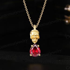 Puede incluir: Un collar dorado con un colgante de calavera y una gema roja en forma de lágrima. La cadena es fina y la calavera tiene detalles. El colgante cuelga de una cadena fina, creando un contraste llamativo con el fondo oscuro.