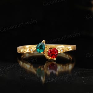 May include: A gold ring featuring a teardrop-shaped emerald green gemstone and a heart-shaped red gemstone. The ring has a crescent moon design and small diamond accents. The words "Customurjewelry" are visible.