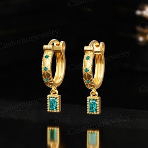 May include: A pair of gold hoop earrings featuring emerald green rectangular gemstones. The earrings have a decorative pattern with small green stones. The text "Customurjewelry" is visible. The earrings are displayed against a dark background.