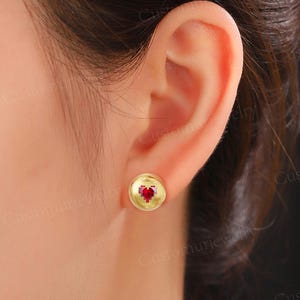 May include: A gold stud earring with a red heart-shaped gemstone is shown close-up. The round earring is set against the ear of a person with brown hair. The background is blurred.
