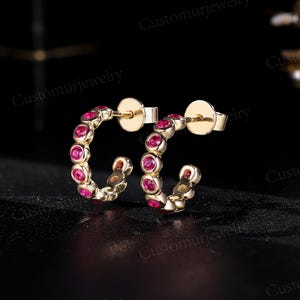 May include: A pair of gold hoop earrings with vibrant red gemstones. The earrings are in a half-circle design, with each earring featuring multiple round, red stones set closely together. The earrings are displayed against a dark background, highlighting their color and design.
