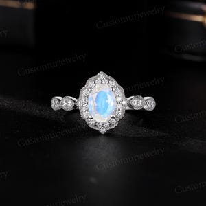 May include: A silver ring featuring an oval, iridescent gemstone, surrounded by a halo of smaller stones. The band has a delicate, detailed design. The ring is set against a dark background, highlighting its sparkle and intricate details. Customurjewelry is printed on the image.