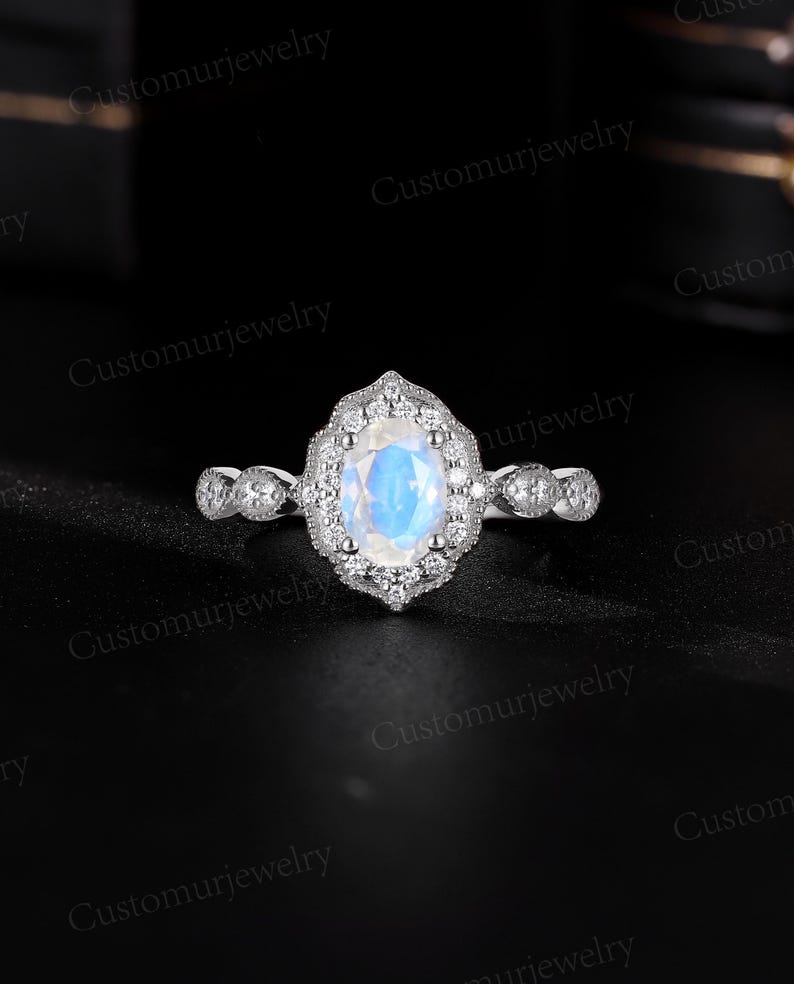 May include: A silver ring featuring an oval, iridescent gemstone, surrounded by a halo of smaller stones. The band has a delicate, detailed design. The ring is set against a dark background, highlighting its sparkle and intricate details. Customurjewelry is printed on the image.