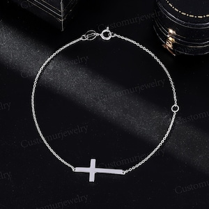 May include: A delicate silver chain bracelet with a simple cross pendant. The chain is thin and delicate, and the cross is small and understated.