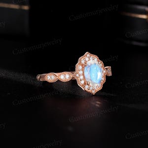 May include: Rose gold ring featuring a large, oval, iridescent gemstone surrounded by a halo of smaller, clear stones. The band has a delicate design with additional small stones. The ring is set against a dark background.