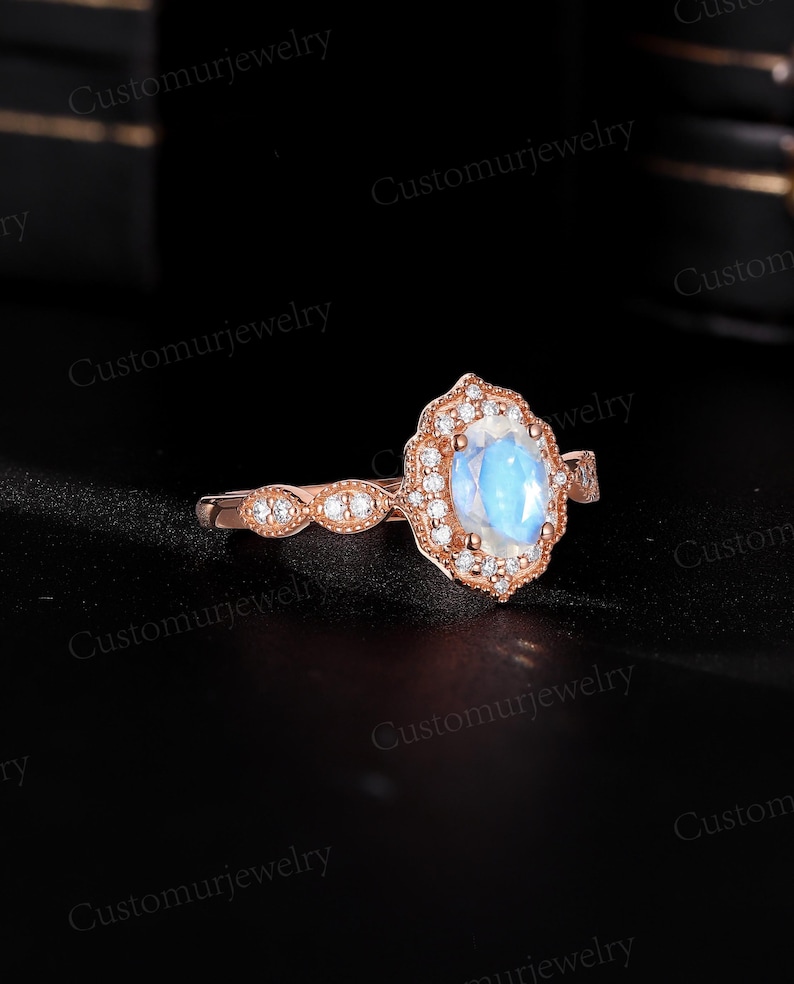 May include: Rose gold ring featuring a large, oval, iridescent gemstone surrounded by a halo of smaller, clear stones. The band has a delicate design with additional small stones. The ring is set against a dark background.