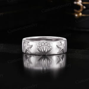 A silver ring with a seashell and starfish design, placed on a reflective surface against a dark background.