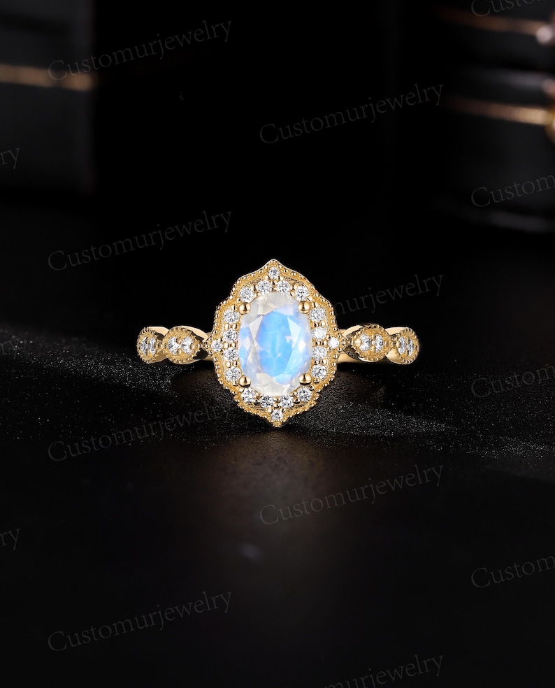 May include: A gold ring with a large, oval, iridescent gemstone at the center, surrounded by smaller clear stones. The band features delicate detailing and additional small stones. The ring is set against a black background.
