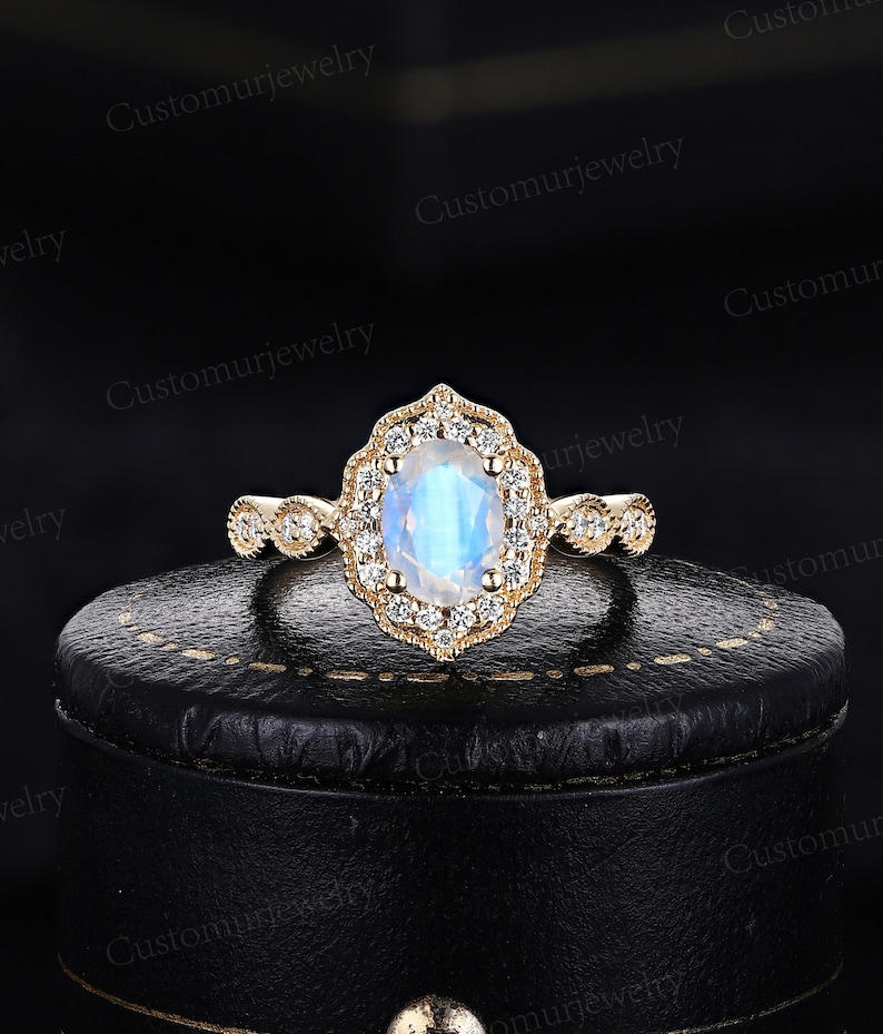 May include: A gold ring with an oval, iridescent moonstone center stone, surrounded by a halo of small, clear gemstones. The ring's band features intricate detailing. The ring is displayed on a black surface.