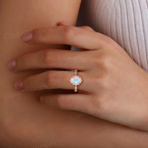 May include: Rose gold ring featuring a light blue oval gemstone, surrounded by a halo of smaller stones. The band has delicate detailing. A piece of jewelry.