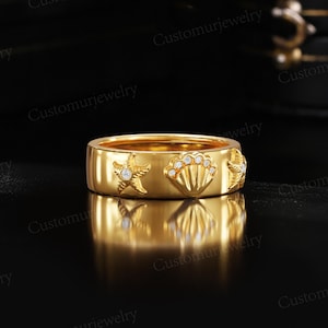 A gold ring with a starfish and seashell design, placed on a reflective surface against a black background.