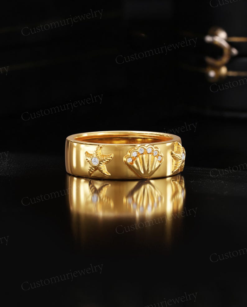 A gold ring with a starfish and seashell design, placed on a reflective surface against a black background.