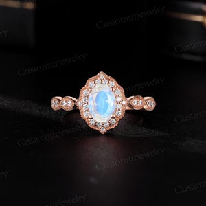 May include: Rose gold ring featuring an oval, iridescent moonstone center stone, surrounded by a halo of small, clear gemstones. The band has a delicate, detailed design with additional small gemstones. The ring is set against a dark background.