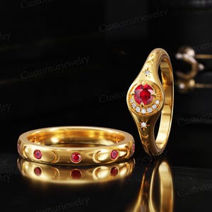 May include: Two gold rings. One ring features a large, round, red gemstone surrounded by small white gemstones and a star design. The other ring is a band with red gemstones and crescent moon cutouts. The rings are on a reflective black surface.