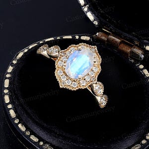 May include: A gold ring with a large, oval, iridescent gemstone, surrounded by smaller clear stones. The ring is set in a decorative, floral-inspired setting. The ring is displayed in a black jewelry box.