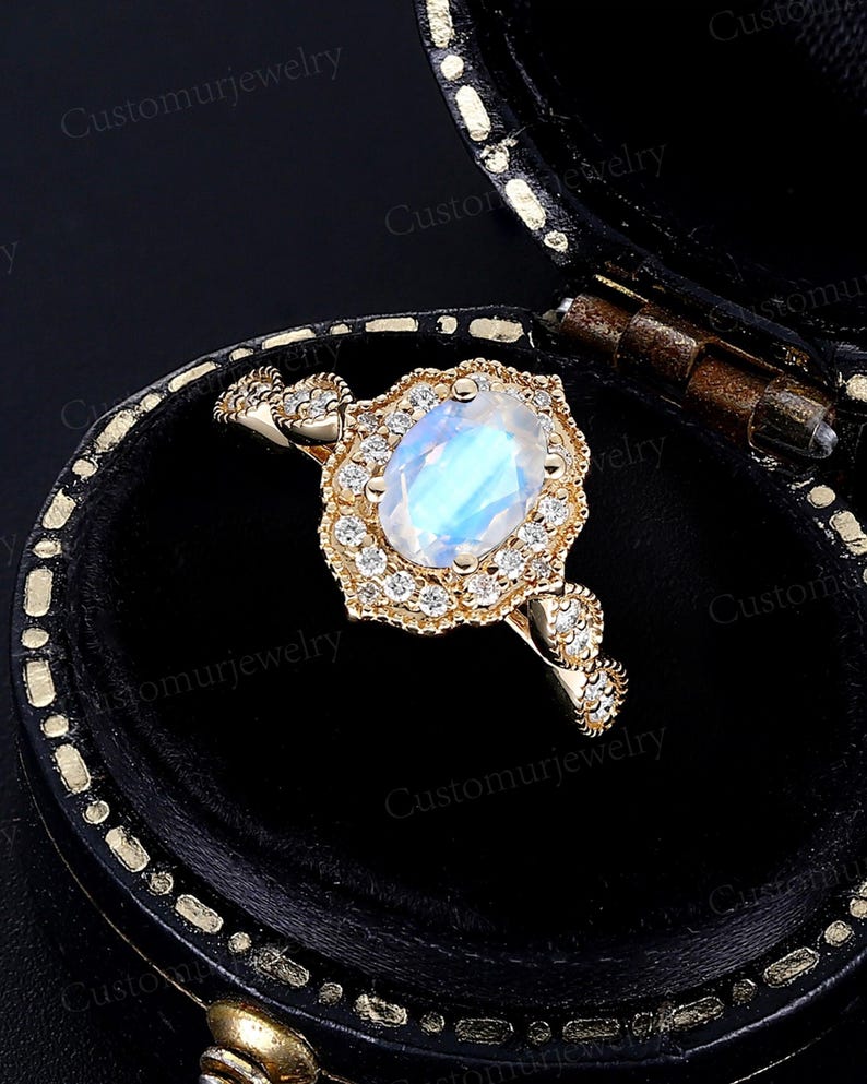 May include: A gold ring with a large, oval, iridescent gemstone, surrounded by smaller clear stones. The ring is set in a decorative, floral-inspired setting. The ring is displayed in a black jewelry box.