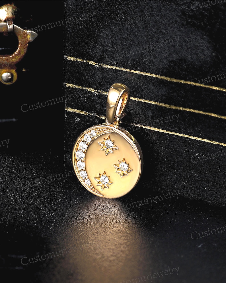 May include: Gold-tone pendant featuring a crescent moon and three stars. The moon is adorned with a row of small, clear gemstones. The stars are also embellished with clear gemstones. The pendant has a loop for a chain.