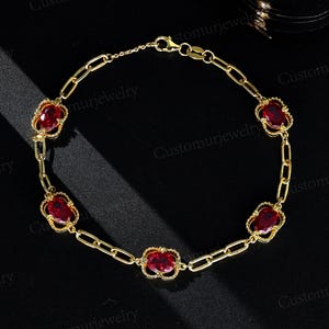 May include: A gold-colored bracelet featuring oval-shaped, deep red gemstones set within ornate, openwork frames. The bracelet is composed of a chain-link design with the gemstones evenly spaced along its length. The clasp is visible at the top.