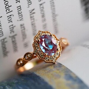 May include: Rose gold ring featuring an oval, color-changing gemstone with a halo of small diamonds. The ring's band has a delicate, ornate design. The gemstone appears to shift between blue and purple hues. The ring is set against a blurred background of text.
