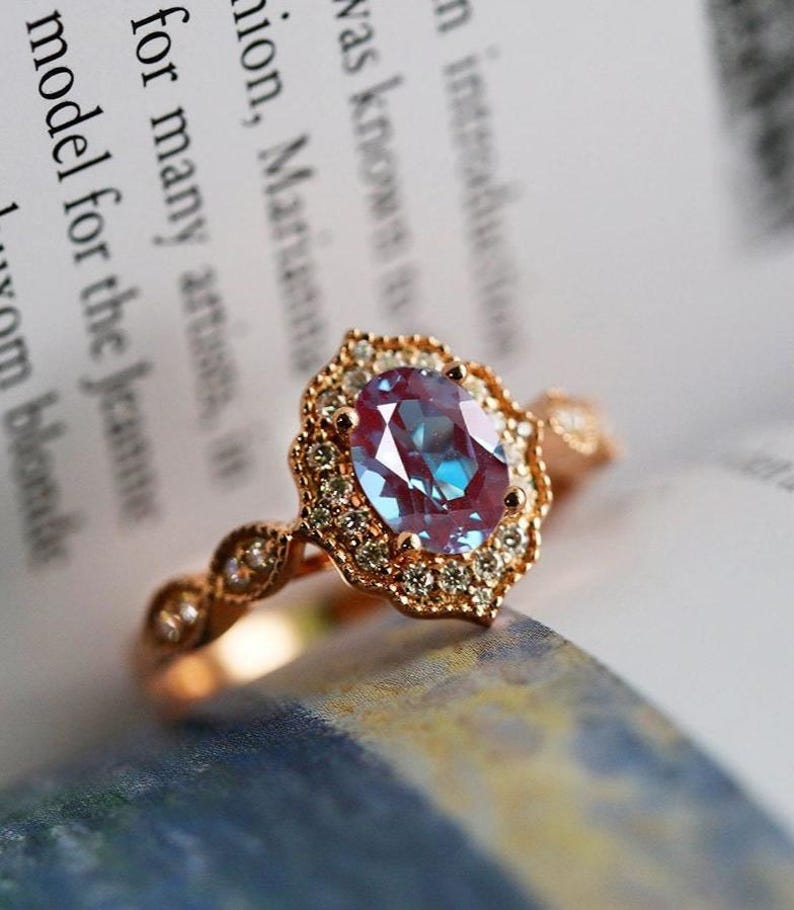 May include: Rose gold ring featuring an oval, color-changing gemstone with a halo of small diamonds. The ring's band has a delicate, ornate design. The gemstone appears to shift between blue and purple hues. The ring is set against a blurred background of text.