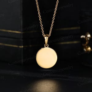 A gold-colored necklace with a round pendant is displayed against a black background.
