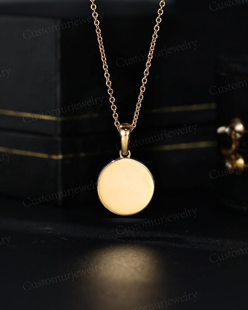 A gold-colored necklace with a round pendant is displayed against a black background.