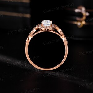 May include: Rose gold engagement ring with a central, round, light-colored gemstone. The ring features a halo of smaller stones around the center and along the band, with intricate detailing. The ring is set against a dark background.