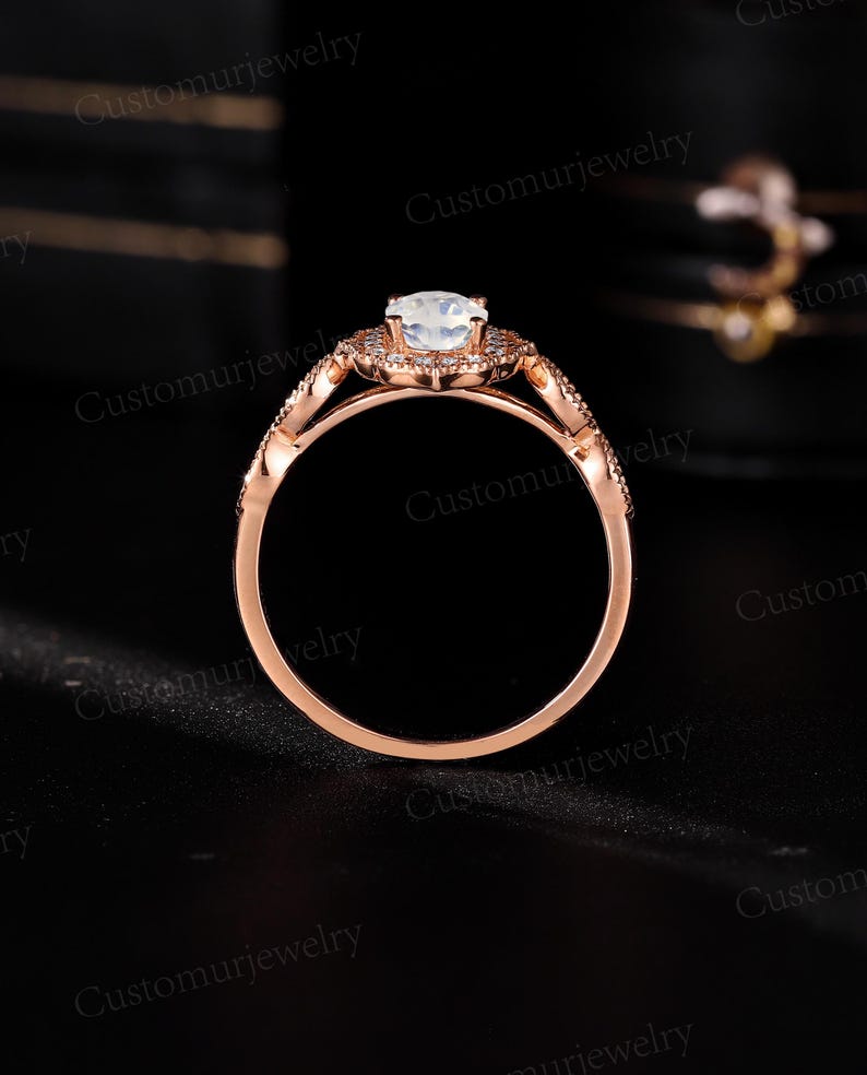 May include: Rose gold engagement ring with a central, round, light-colored gemstone. The ring features a halo of smaller stones around the center and along the band, with intricate detailing. The ring is set against a dark background.