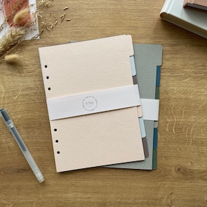 Könnte beinhalten: Ein Satz von fünf Trennern für einen Planer oder ein Notizbuch. Die Trenner sind in verschiedenen Grautönen und Blau. Die Trenner sind mit dem Text "d'Anni" in einem weißen Kreis beschriftet.