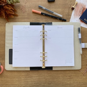 Könnte beinhalten: Geöffneter beiger Planer mit goldenen Ringen, zeigt die Seiten für Dezember und Januar. Der Planer hat einen schwarzen Rücken und einen kleinen Kalender. Stifte, Büroklammern und andere Schreibwaren liegen auf Holz.