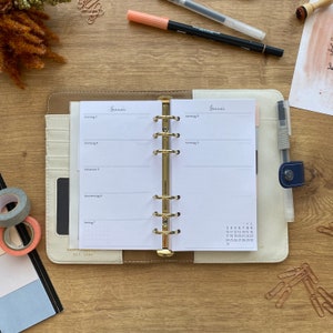 Könnte beinhalten: Geöffneter Planer mit goldenen Ringen, mit Wochenkalenderseiten mit deutschem Text. Der Planer ist cremefarben und beige mit einem marineblauen Stifthalter. Stifte, Washi Tape und Büroklammern auf Holzoberfläche.