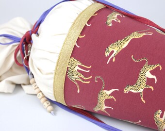 May include: A cone-shaped fabric gift bag featuring a maroon background with a repeating yellow and black cheetah print. The top section is cream-colored fabric gathered with purple and red drawstrings, accented by a gold ribbon and wooden beads.