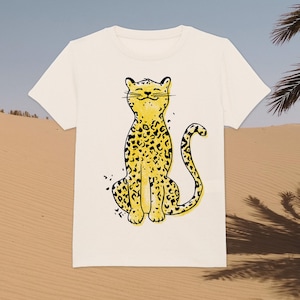 Może przedstawiać: Biała koszulka z kreskówkową ilustracją żółtego lamparta z czarnymi plamami siedzącego. Lampart ma szczęśliwy wyraz twarzy.