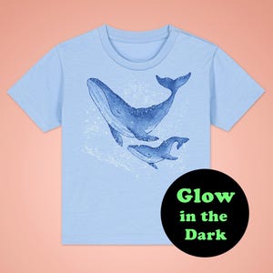 Könnte beinhalten: Hellblaues T-Shirt mit einem im Dunkeln leuchtenden Design von zwei Walen. Der Text "Glow in the Dark" ist auf einem schwarzen Kreis gedruckt.