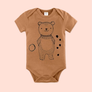 Może przedstawiać: Brązowy, krótkorękawny śpioszek dla niemowląt z czarno-białą ilustracją niedźwiedzia w swetrze. Niedźwiedź stoi na brązowym tle z czarnymi kropkami i zielonym kółkiem.
